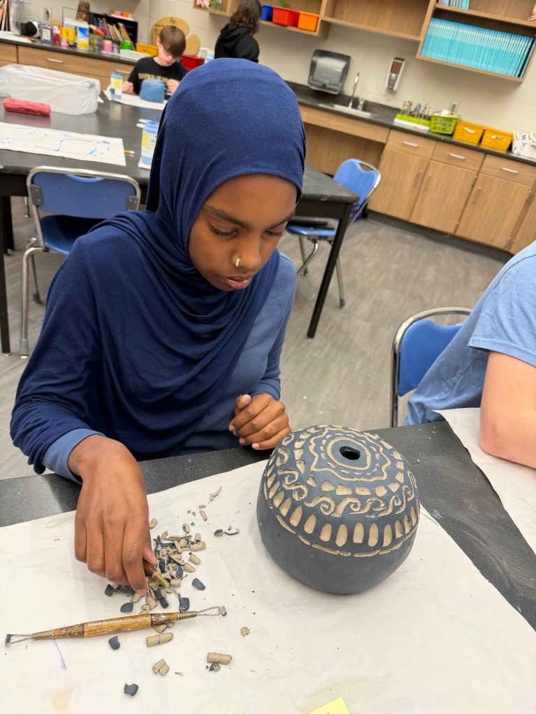 A young girl in a blue hijab is carefully working on a decorated ceramic pot, surrounded by small pieces of clay. She is using a sculpting tool while focusing on her craft in an art classroom.