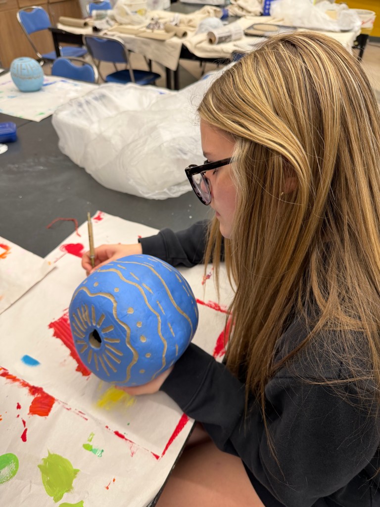 A young girl with long hair is painting a blue and gold decorated pumpkin in an art classroom. She is focused on her work with a paintbrush in hand.