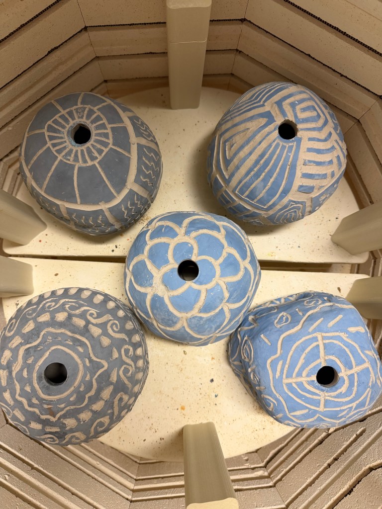 Four decorated ceramic pieces in shades of blue, featuring various patterns, placed inside a kiln for firing.