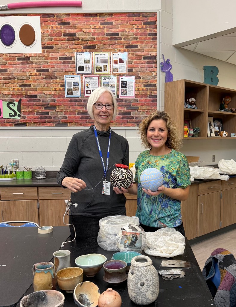 Two women holding ceramic pots in an art classroom, with various pottery pieces displayed on a table and an educational bulletin board in the background.