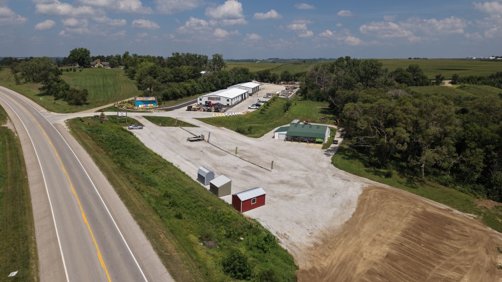 Aerial view of a vacant lot with a gravel surface and several small buildings, including a red shed. Nearby, a warehouse building and open fields, with a road visible on the left.