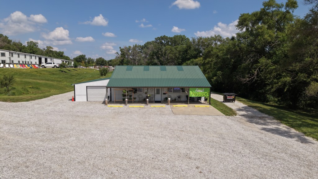 Aerial view of a green-roofed produce store with a large sign reading 'Fresh Produce' and a gravel parking area, surrounded by greenery and a nearby white building.