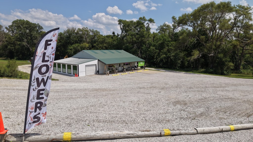 A flower shop with a green metal roof and large windows, surrounded by gravel. A flag reading 'Flowers' is visible in the foreground, with trees in the background under a blue sky.