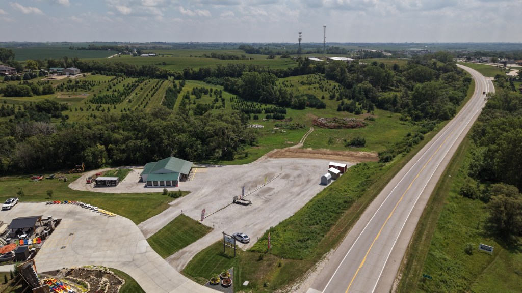Aerial view of a rural landscape featuring a newly developed commercial area with a small green-roofed building, gravel parking lot, and adjacent highway. In the background, there are rows of trees and agricultural fields.