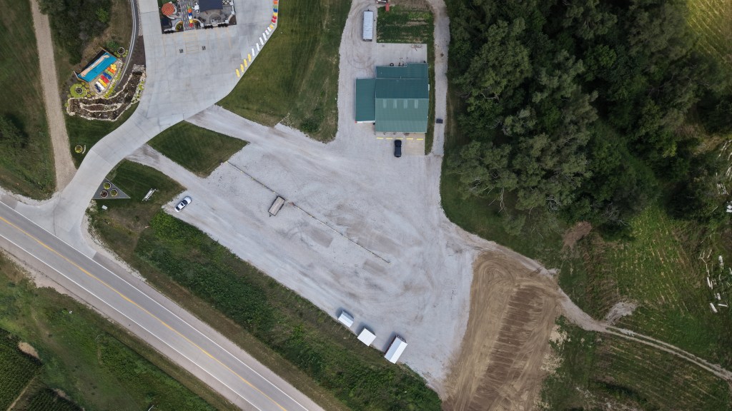 Aerial view of a gravel parking lot with a building and vehicles, surrounded by greenery and a road.