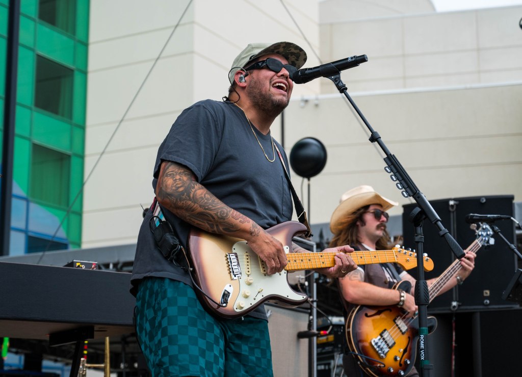 A musician wearing sunglasses and a cap performs on stage, playing an electric guitar while singing into a microphone. Another band member plays the bass guitar in the background.