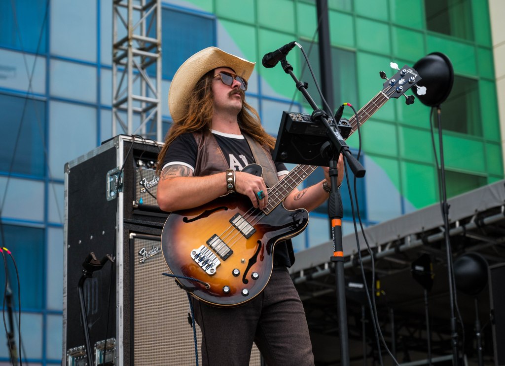 A musician playing an electric bass guitar on stage, wearing a cowboy hat and sunglasses, with long hair and a rock band outfit, in front of a colorful backdrop of a building.