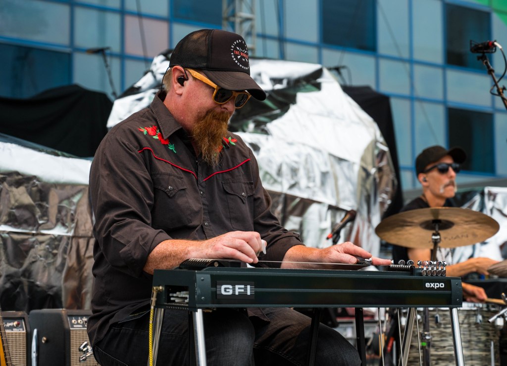 A musician playing a steel guitar on stage, wearing a black shirt with red embroidery and a baseball cap. In the background, a drummer is visible.