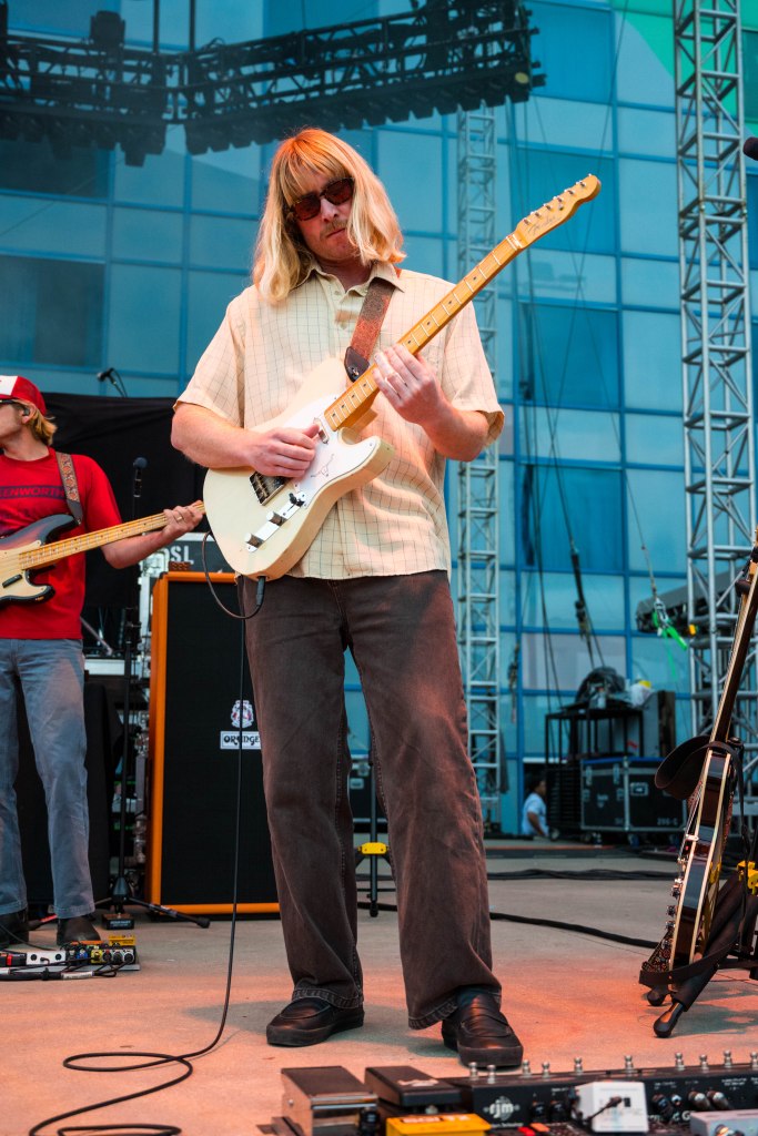 A guitarist performing on stage, wearing sunglasses and a light-colored shirt, playing an electric guitar. A band member is visible in the background, and various music equipment is set up around them.