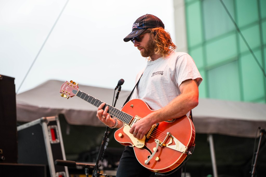 A musician playing an orange Gretsch guitar on stage, wearing a gray t-shirt and baseball cap.