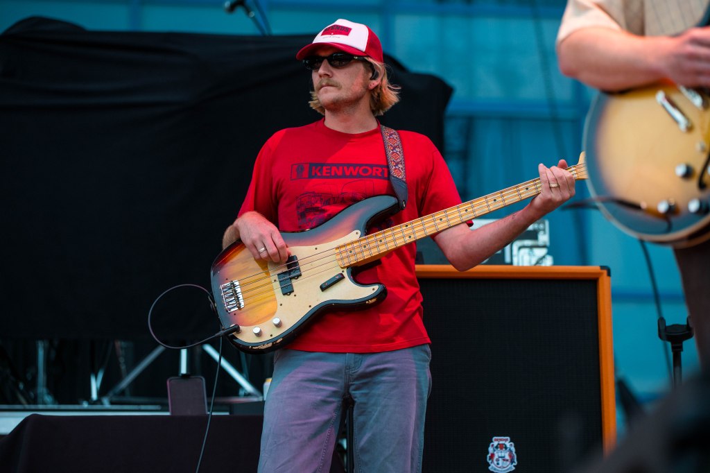 A musician playing a bass guitar on stage, wearing a red t-shirt and a white cap with sunglasses, with part of an amplifier visible in the background.
