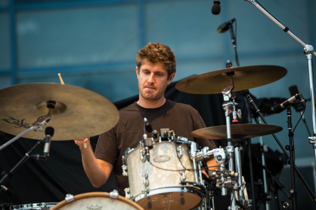 A male drummer performing on stage with a focused expression, wearing a black shirt, surrounded by drum equipment.