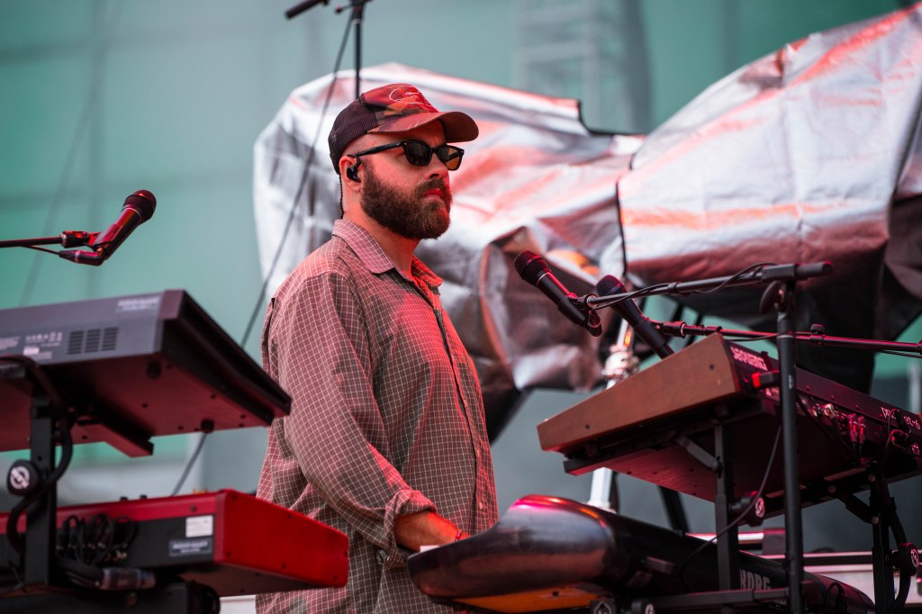 A musician wearing a cap and glasses performs on stage in front of keyboards, with a microphone in the foreground.