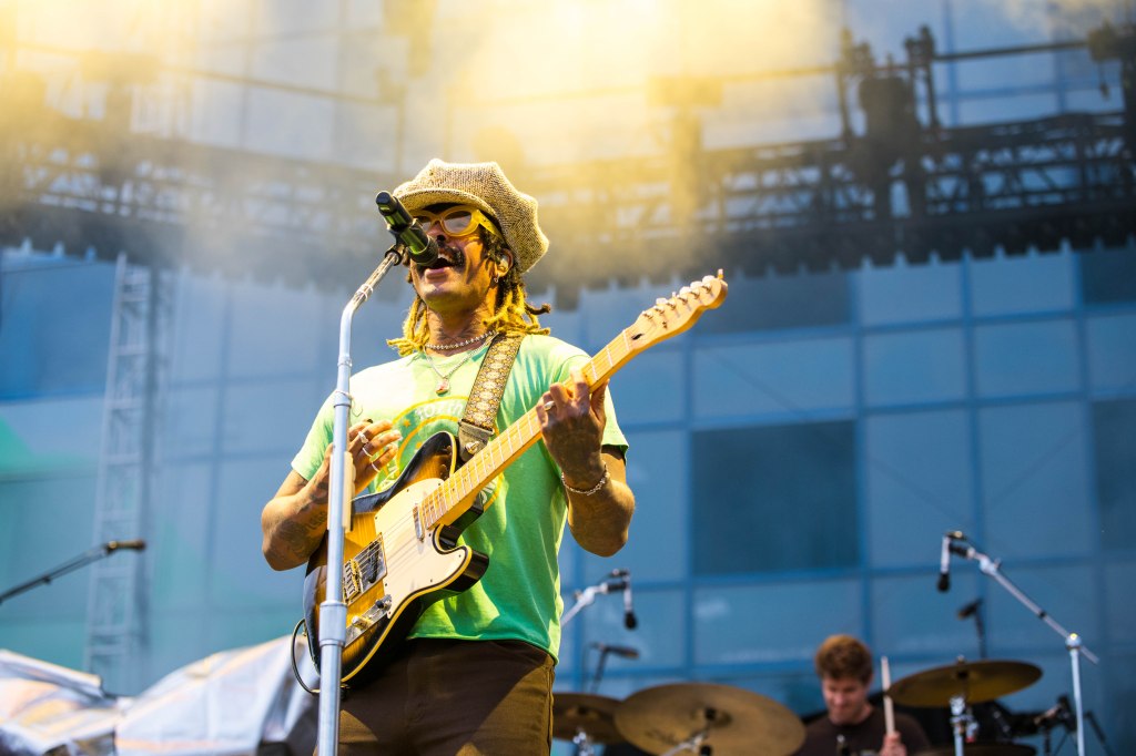 Musician performing on stage with a guitar and microphone during a concert, wearing sunglasses and a hat.