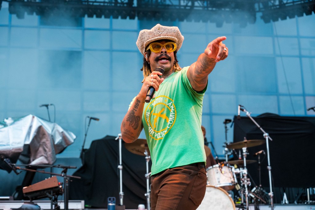 A performer on stage singing into a microphone, wearing a green shirt and a patterned hat, with a lively expression and sunglasses.