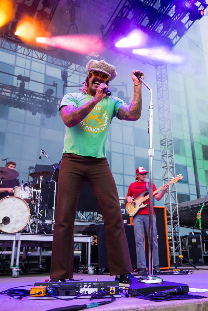 A musician performing on stage, wearing a green shirt and a gray hat, singing into a microphone with energetic body language. Behind him, a drummer and a bassist are visible, along with colorful stage lights.