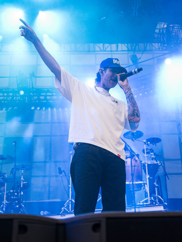 A male singer performs on stage, holding a microphone and pointing with one hand, surrounded by blue stage lights and a smoke effect.