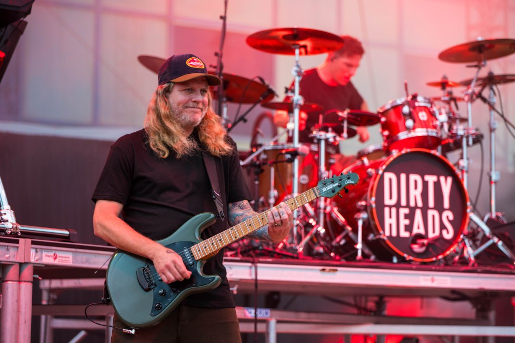A guitarist with long hair playing an electric guitar on stage, with a drummer visible in the background. The band's name 'DIRTY HEADS' is displayed on a drum kit.
