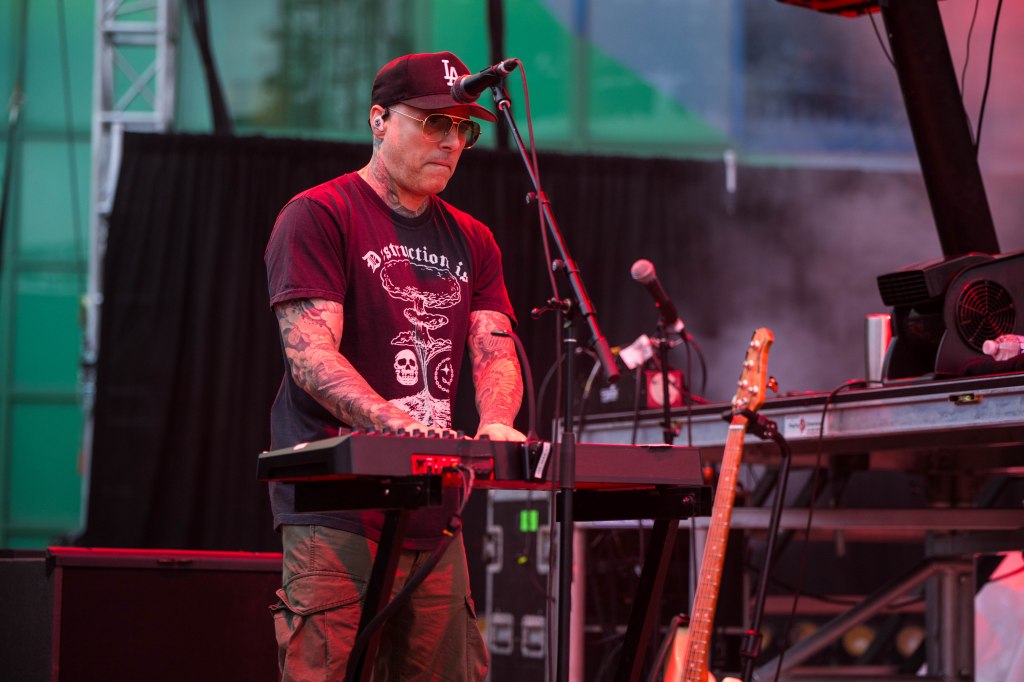 A musician playing a keyboard on stage, wearing sunglasses and a black 'Destruction' t-shirt, with a microphone and guitar in the background.