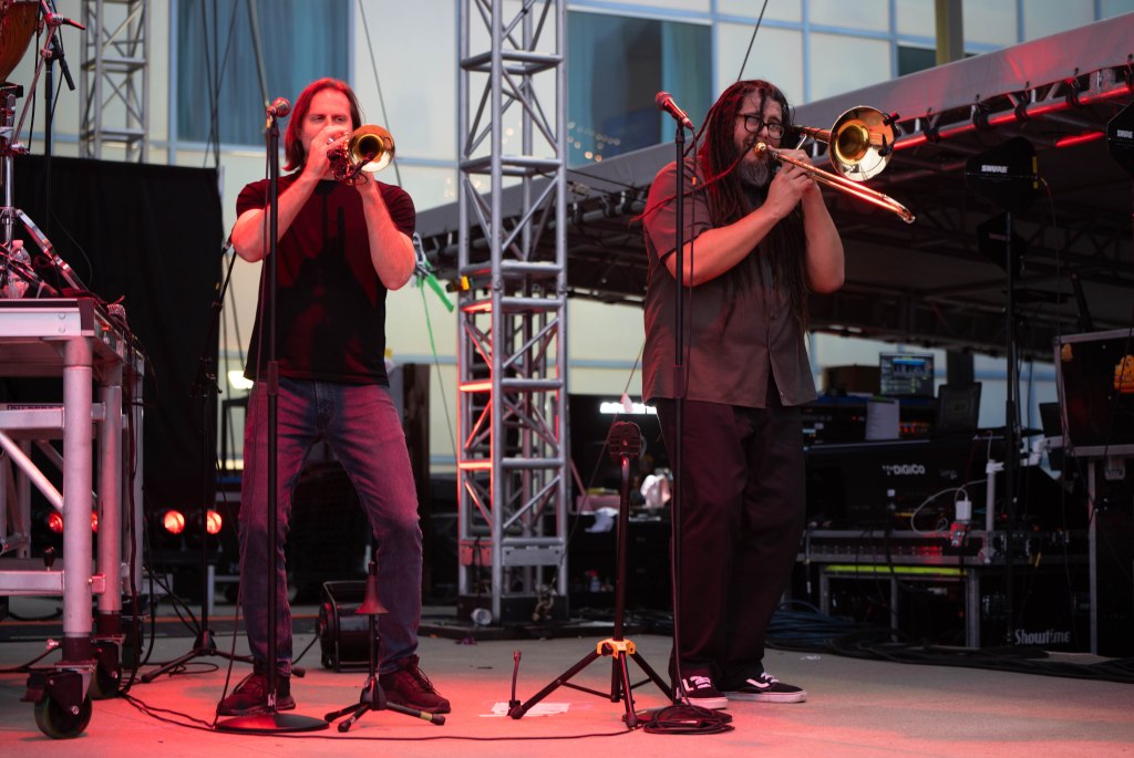 Two musicians playing trombones on stage during a live performance, with equipment and lighting in the background.