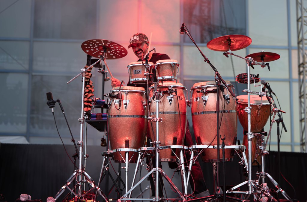 A drummer performing on stage with a set of conga drums and cymbals, surrounded by a misty atmosphere.