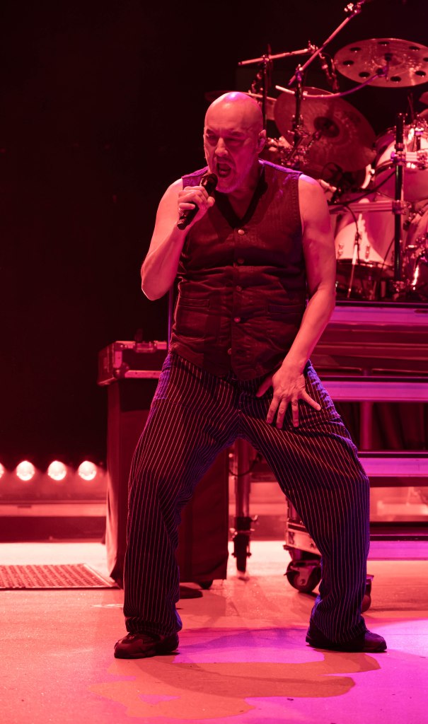 A performer singing passionately on stage, wearing a sleeveless shirt and striped pants, with dramatic lighting in the background.