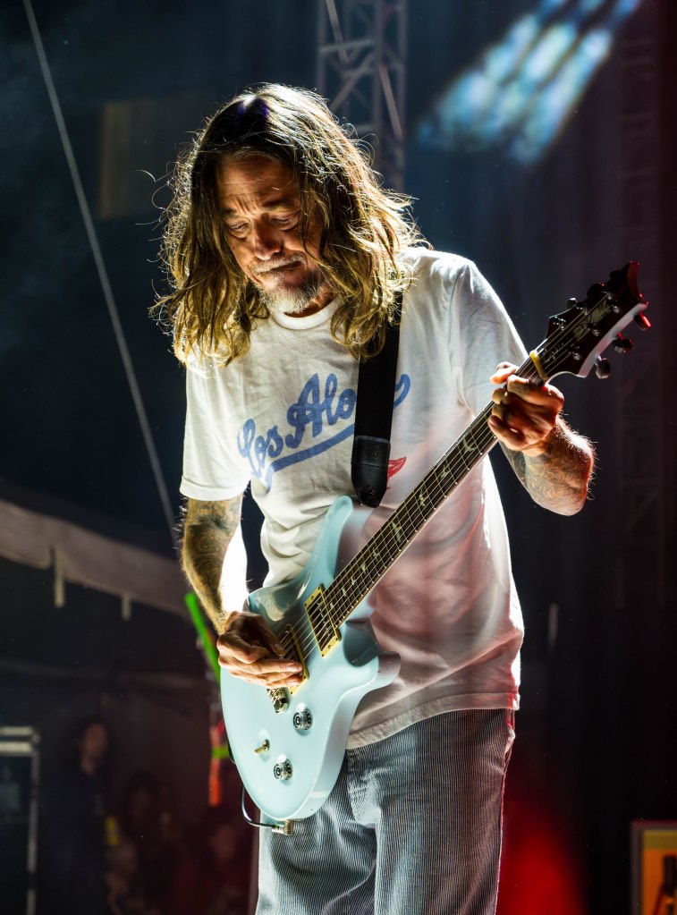 A musician performing on stage, wearing a white t-shirt and blue striped pants, playing a light blue guitar with long hair and a focused expression.