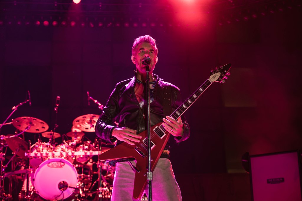 A musician performing on stage with a guitar, wearing a leather jacket, under pink stage lights with a drum set in the background.