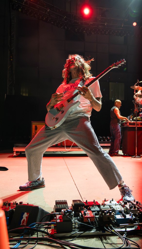 A guitarist performing energetically on stage, dressed in casual clothing and surrounded by musical equipment and lighting.