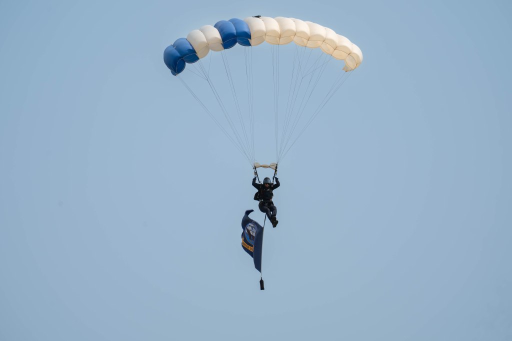 A parachutist in a black jumpsuit descends from the sky, holding a flag, with a blue and white parachute above.