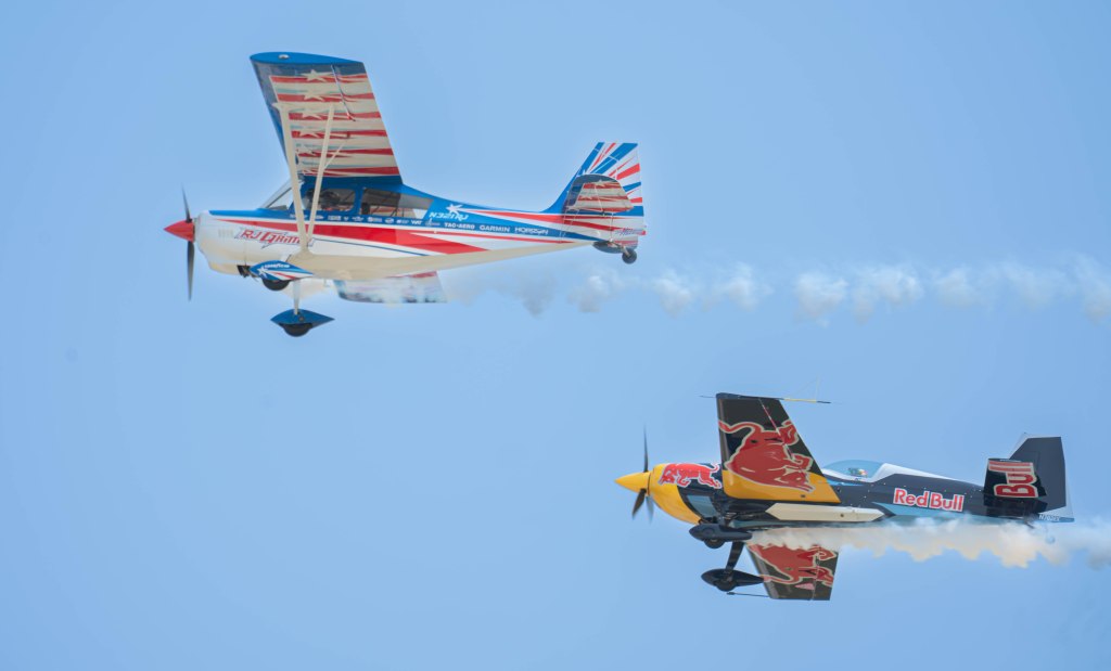 Two airplanes performing aerial maneuvers against a clear blue sky, one adorned with red, white, and blue stripes and the other featuring a Red Bull logo, with smoke trailing behind them.