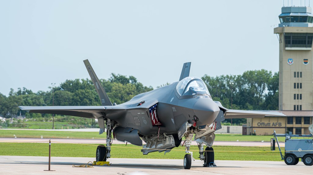A modern fighter jet on the tarmac at an airfield, with a control tower in the background.