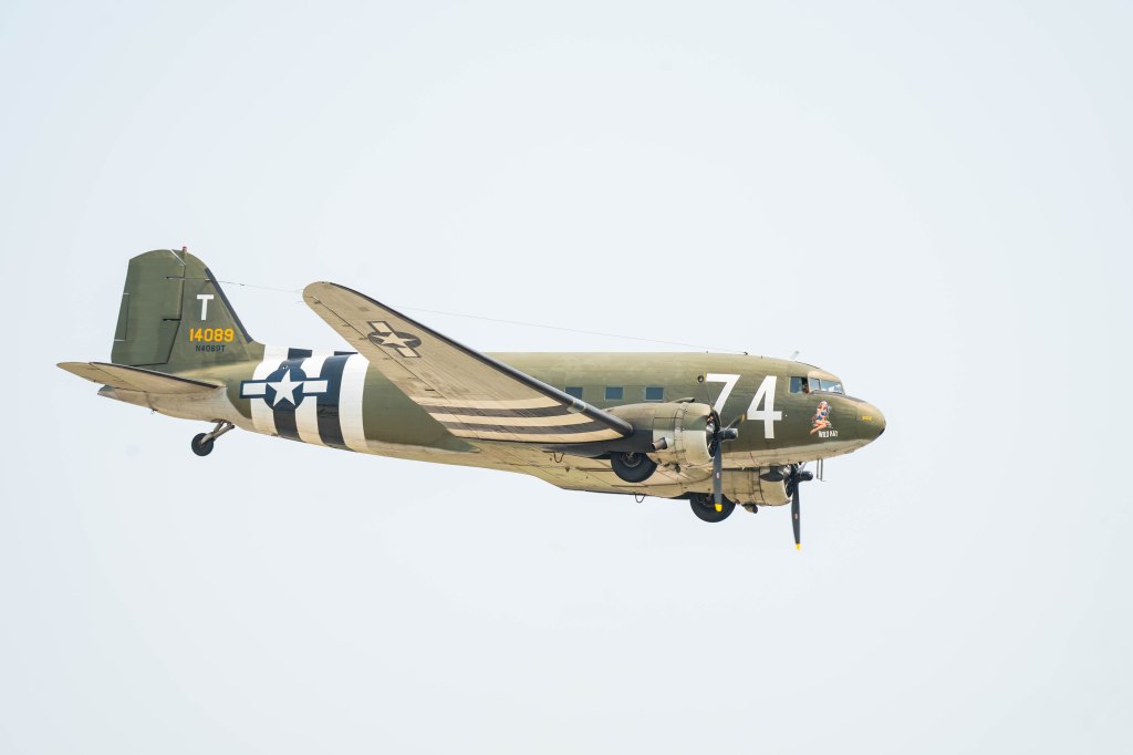 A vintage military aircraft flying in the sky, featuring olive green and white stripes, with identification numbers and a painted character on the nose.