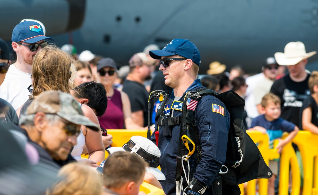 A crowd of people gathered at an event, with a military personnel in a blue jumpsuit and helmet standing in the foreground, wearing a parachute harness.