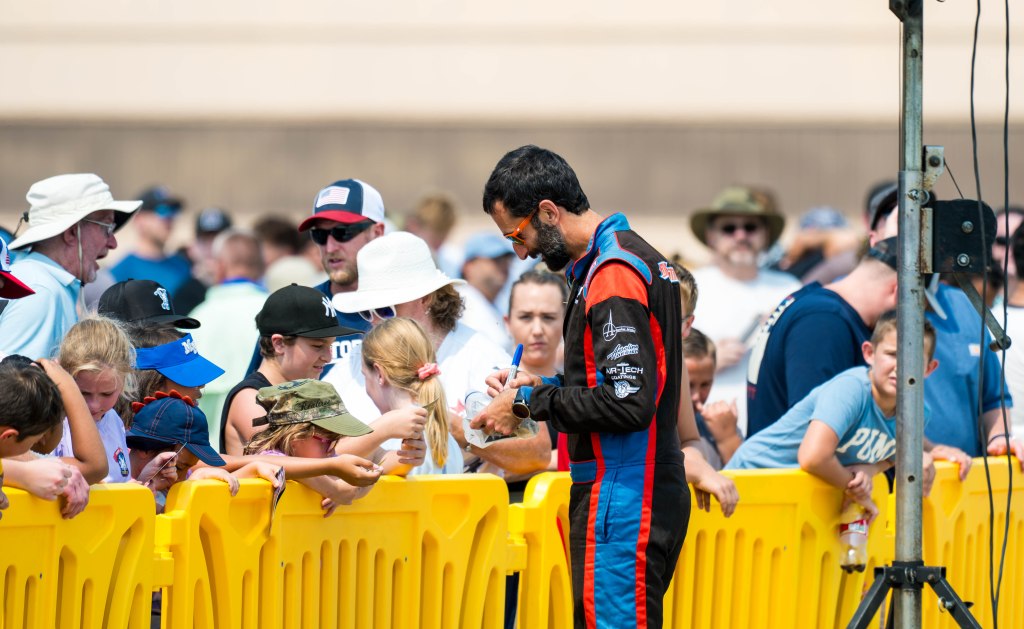 A racecar driver interacts with a crowd of fans, including children, at an event, signing autographs in front of a yellow barrier.