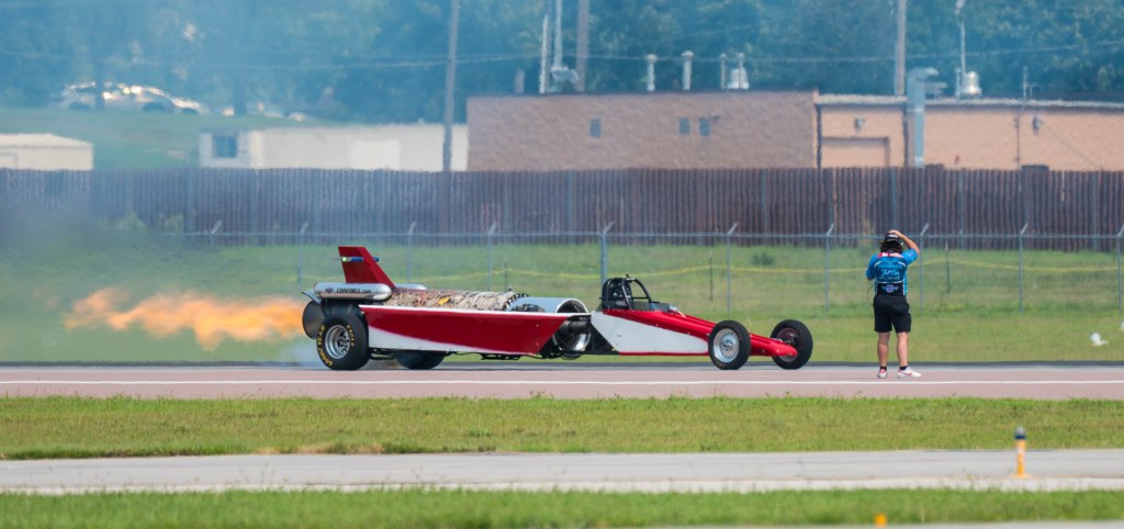 A drag racing car with flames shooting from the exhaust, on a runway, while a person in a blue shirt waves nearby.