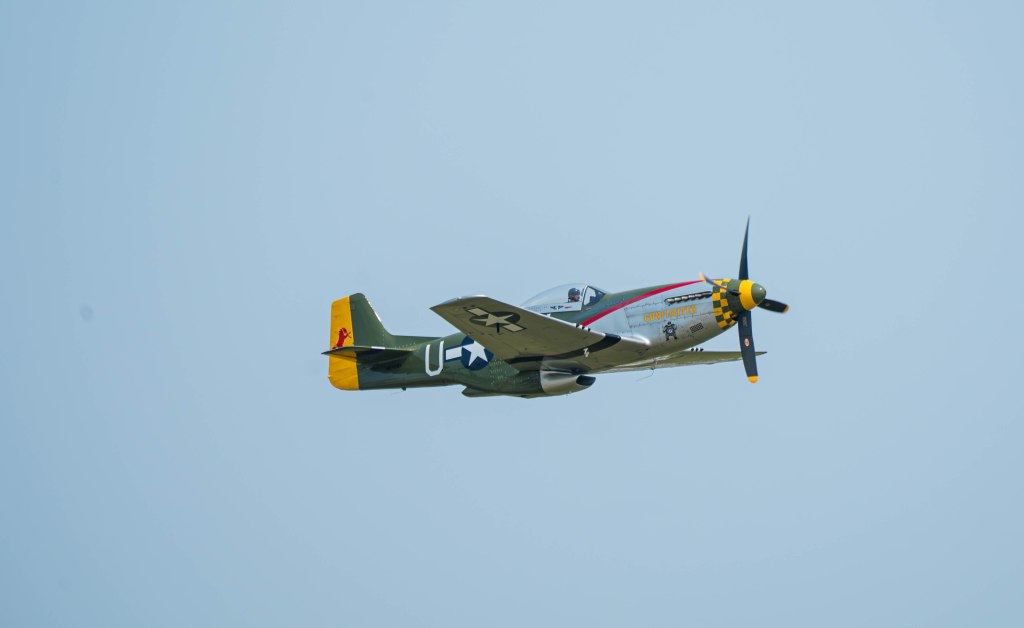 A vintage military aircraft flying against a clear blue sky, showcasing a green and yellow paint scheme with distinctive markings.