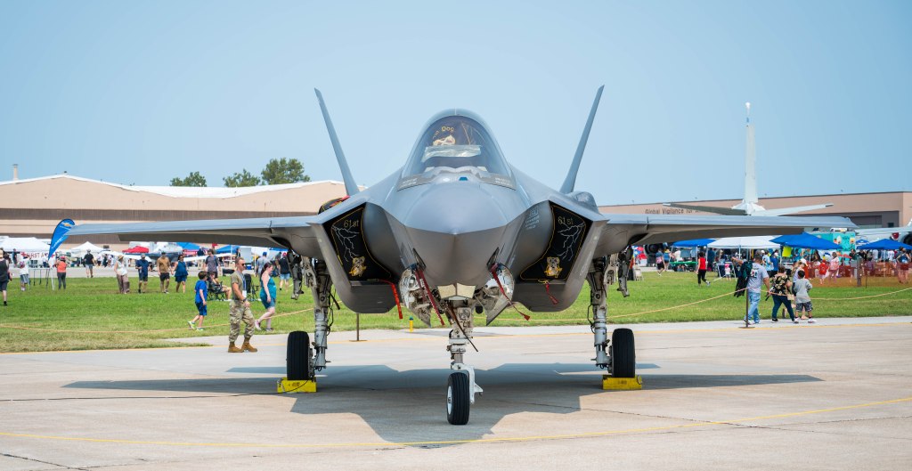 Front view of a military jet on display at an airshow, with spectators in the background.