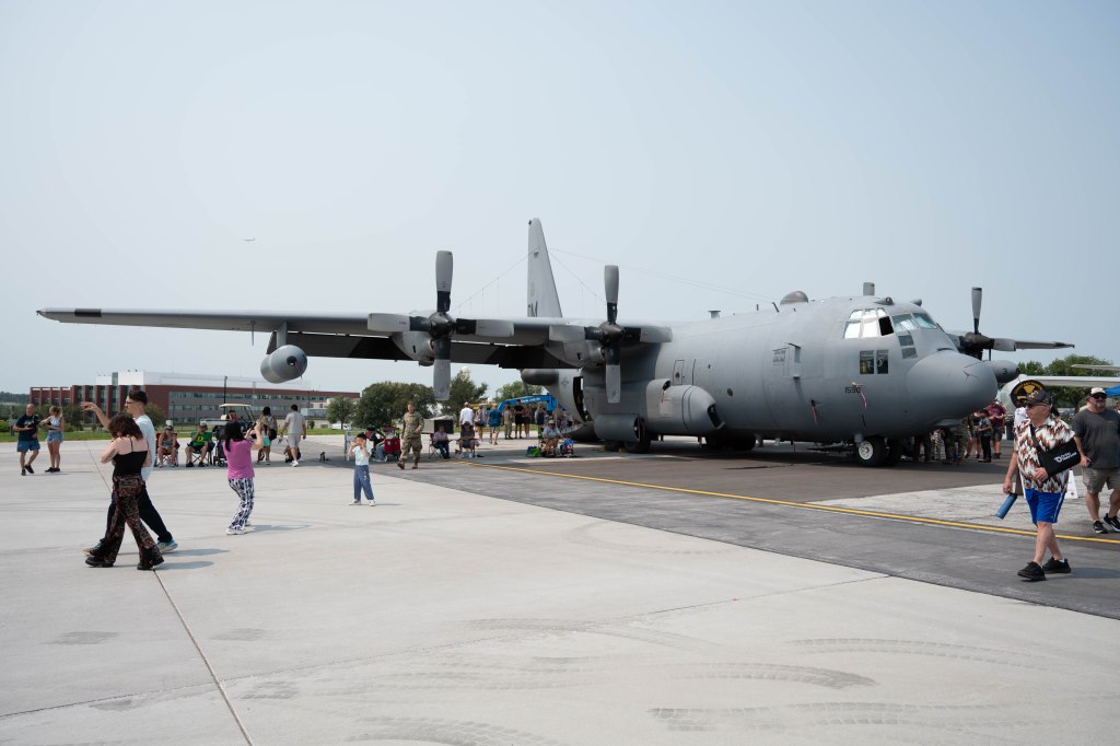 A large military aircraft parked on a concrete tarmac, with a crowd of people walking around and viewing the plane.