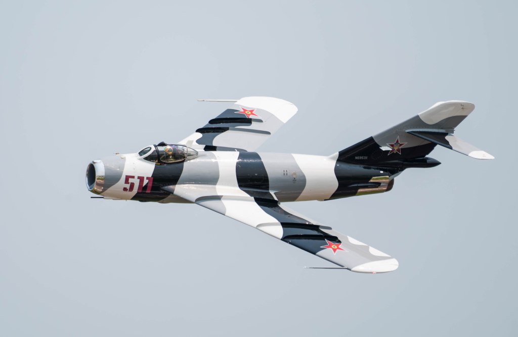 A military jet in flight, featuring a distinctive gray and white camouflage pattern with a red star emblem.