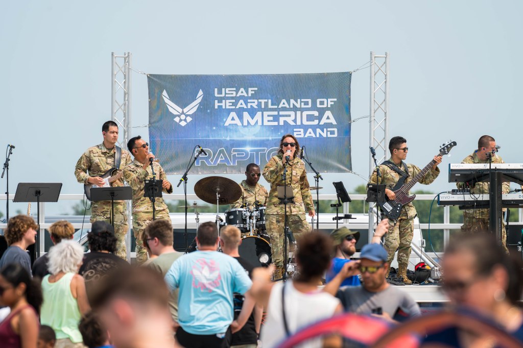 A military band performing on stage at an outdoor event, with members in uniform playing various instruments, including guitar, bass, and drums, in front of an audience.