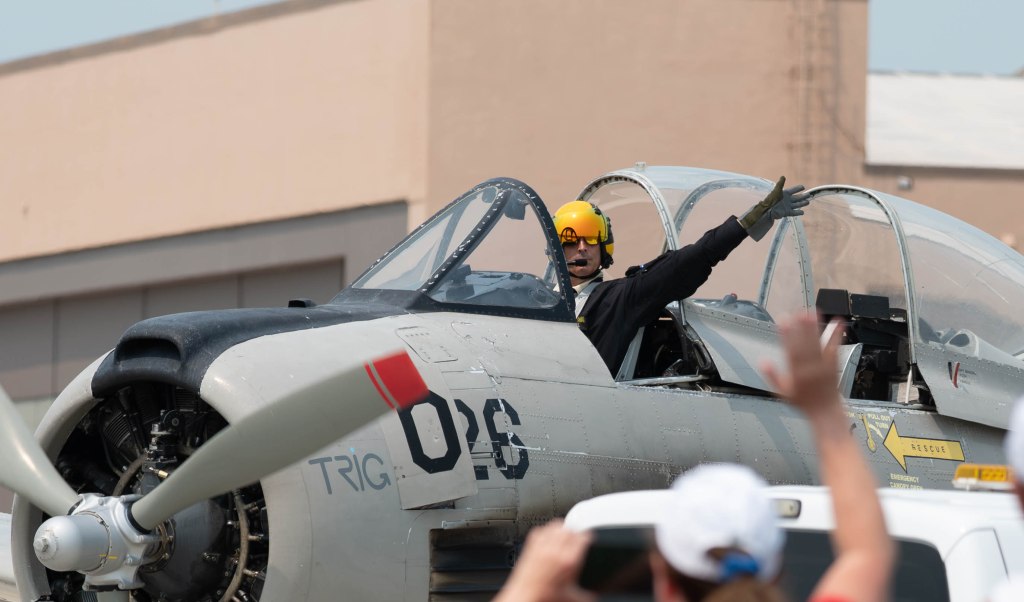 Pilot in a vintage aircraft cockpit giving a salute, wearing a yellow helmet and gloves, with people waving in the foreground.