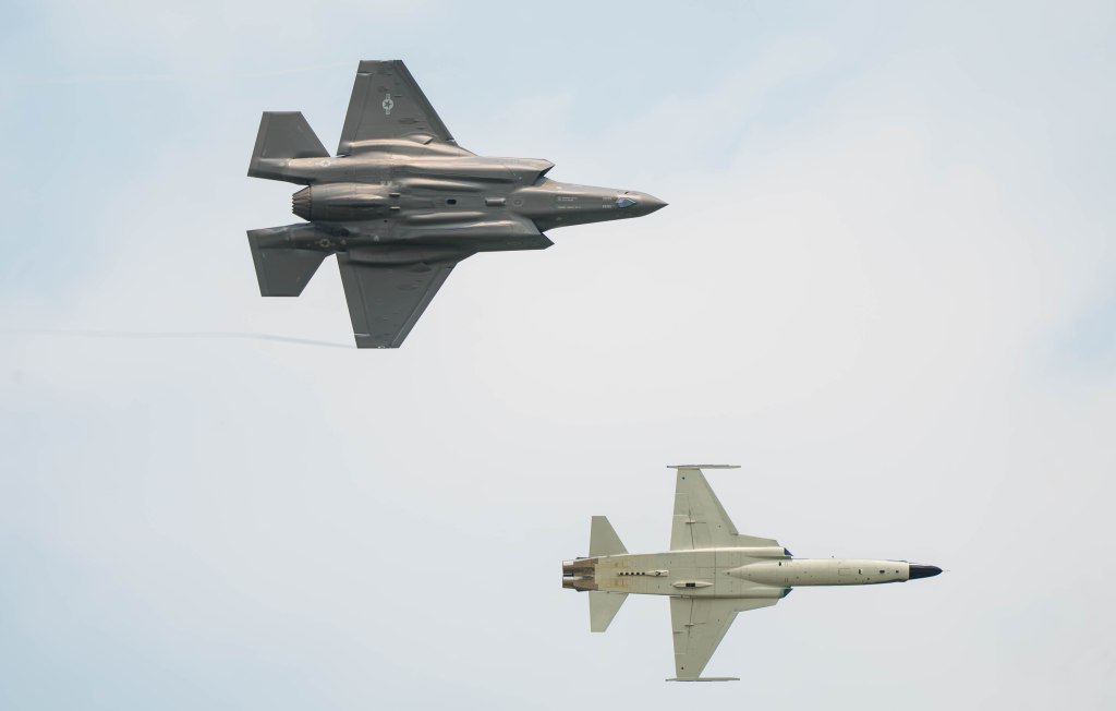 An F-35 fighter jet flying in formation with an unmanned aerial vehicle against a cloudy sky.