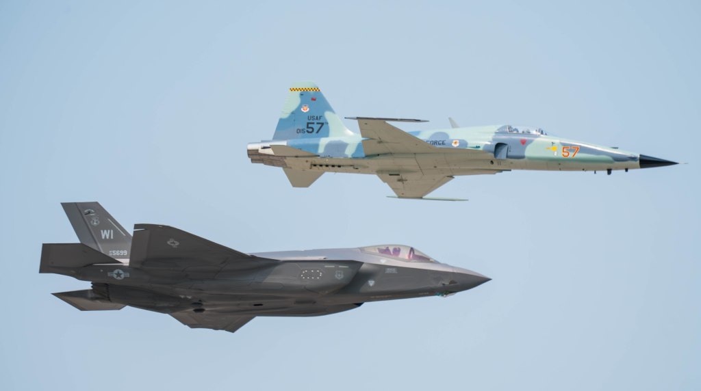 An F-35 fighter jet flying below an F-16 fighter jet in a clear blue sky.