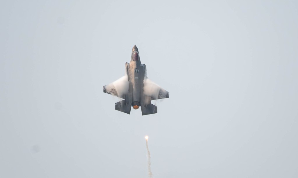 A fighter jet ascending vertically, emitting smoke from its engines, with a missile being launched from its underside.