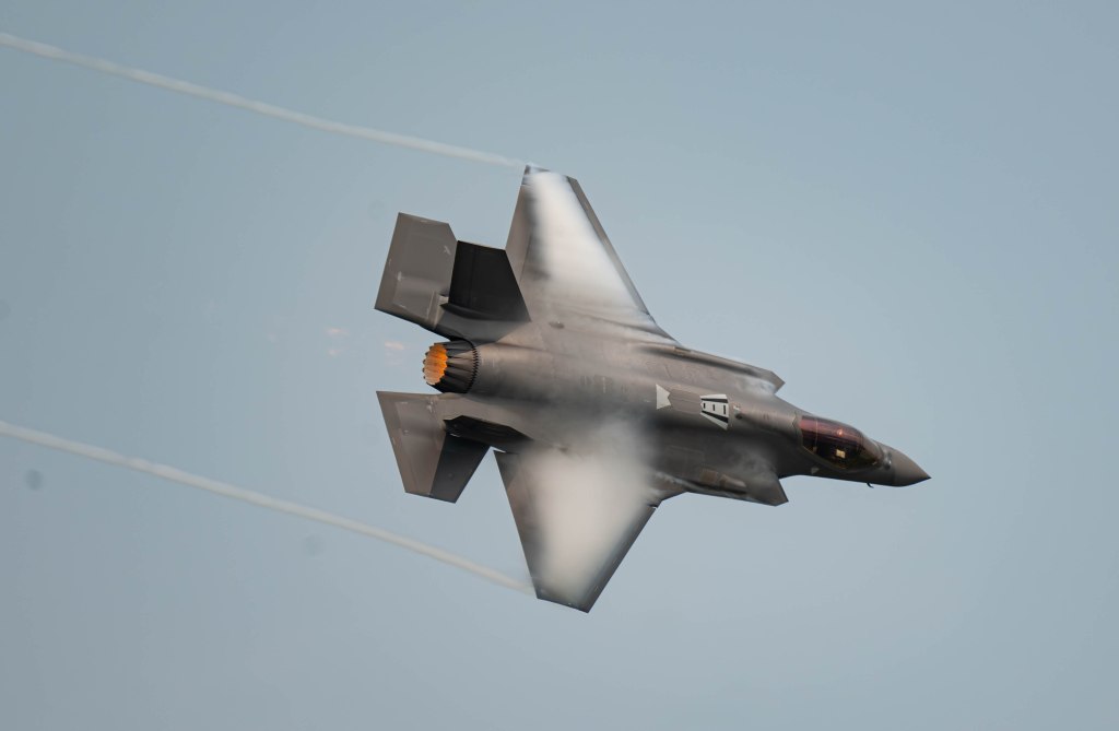 A military jet performing a high-speed maneuver, showing vapor trails and flames from its engine.
