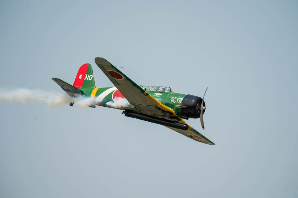 A vintage aircraft performing a high aerial maneuver, with green and red markings and smoke trailing from its wings.