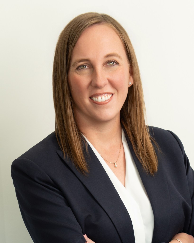 A professional headshot of a woman with long brown hair, smiling confidently while wearing a dark blazer over a white top.