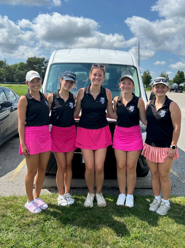 Five young women posing together in athletic attire, consisting of black tops and pink skirts, in front of a van on a sunny day.