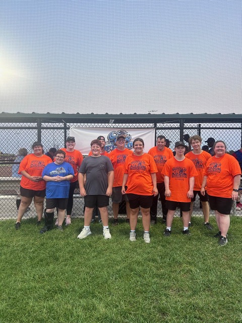 A group of young athletes wearing matching orange t-shirts poses together on a grassy field, with a fence and a banner in the background.